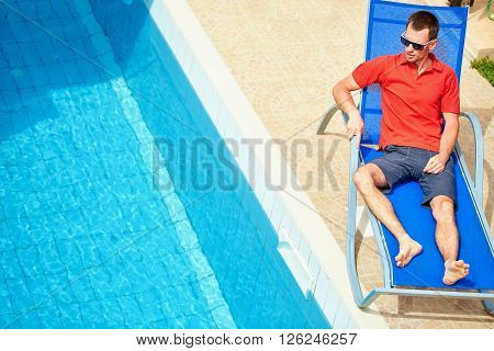 young man relaxing on the chaise-longue near the pool. man wearing in red polo shirt and sunglass