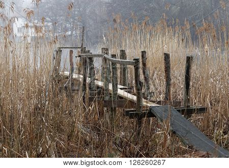 Rural view of old bridge leading through the reed to the lake. Houses in the background