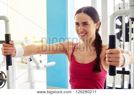 Young Caucasian smiling girl in sportswear is working out on butterfly machine in gym
