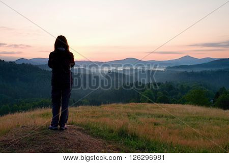 Long hair woman stand on meadow with golden stalks of grass and watch over misty and foggy morning valley to sunrise