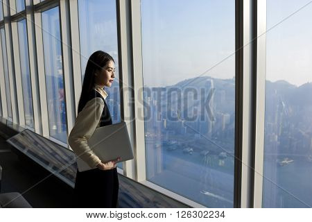 Asian skilled businesswoman is holding laptop computer while is standing in modern office interior near big window with cityscape view. Female executive looking satisfied after her successful meeting