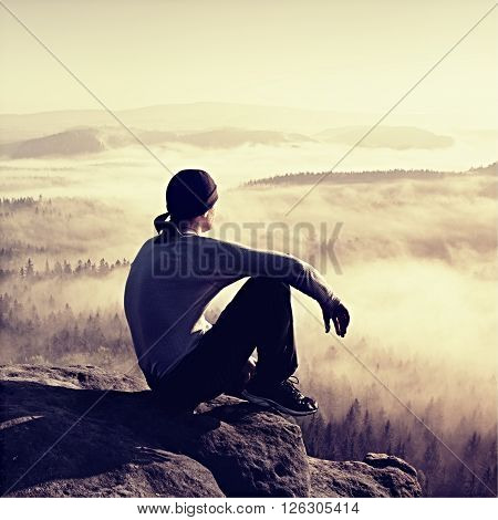 Man sit on the peak of rock and watching into colorful mist and fog in forest valley.
