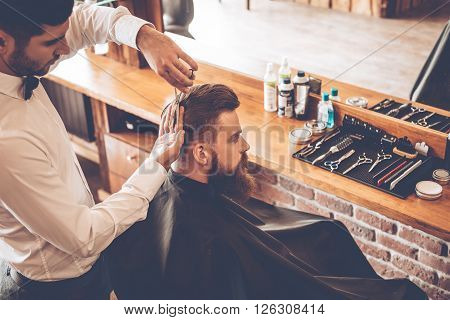 Haircut must be perfect. Top view of young bearded man getting haircut by hairdresser while sitting in chair at barbershop