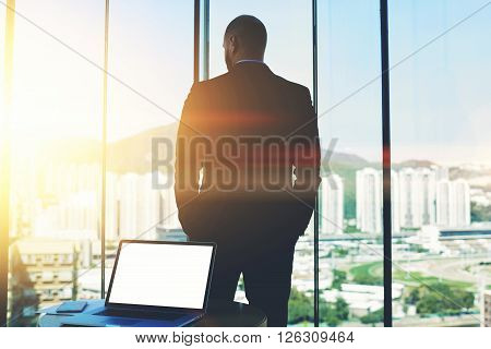 Silhouette of man CEO is thinking about future of his company while is watching in office window on a skyscrapers business center. Open net-book with mock up copy space screen is lying on the table