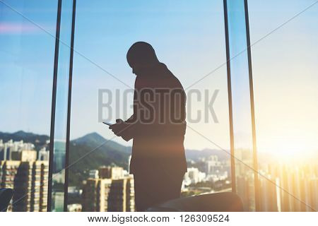Silhouette of a man entrepreneur is typing text message on mobile phone to investor to consider again the question of funding while is standing in office near big skyscraper window with city view