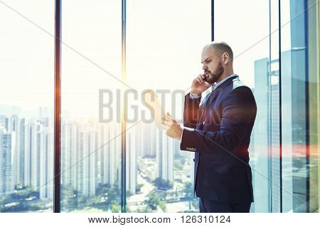 Confident man successful entrepreneur dressed in suit is reading the monthly report on portable digital tablet and talking via cell telephone with manager his company during business trip in Hong Kong