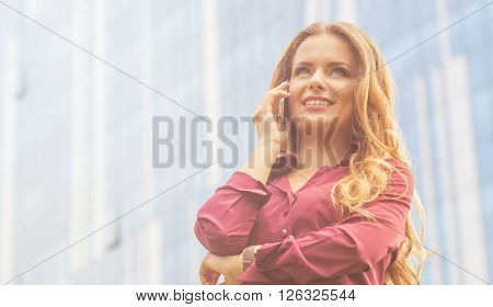 Businesswoman working with her partners in the city over mobile phone. Woman in vinaceous blouse standing near office building. Toned image.