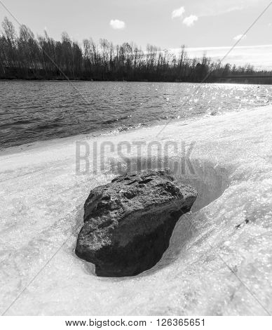 Stone in Ice by River with trees and a patrly cloudy sky