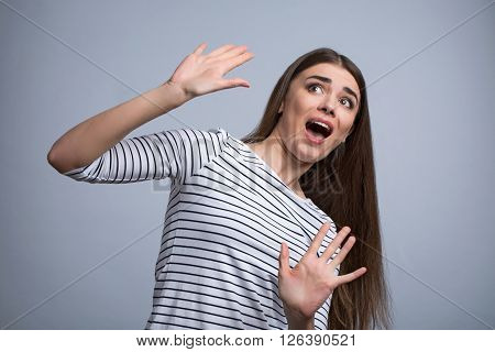 Pleasant scared  beautiful girl holding her hands up   evading danger while standing isolated on grey background