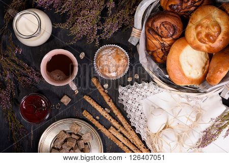 Baked pastries and bread sticks with milk, boiled eggs, cocoa and jam on rustic wooden table. Top view. Decorated with heather.