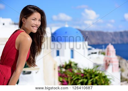 Santorini tourist Thira Greece island tourism - Asian woman wearing red dress on summer travel looking at view with the famous attraction three domes chapel church. Luxury destination.