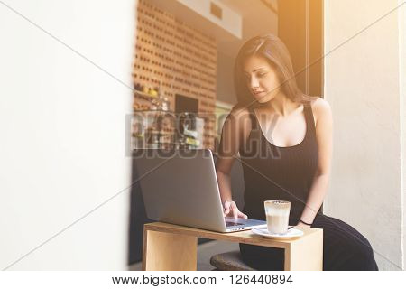 Young female freelancer engage in remote work in internet by using her laptop computer during breakfast in cafe. Gorgeous latin woman connecting to wireless via net-book while breakfast in coffee shop