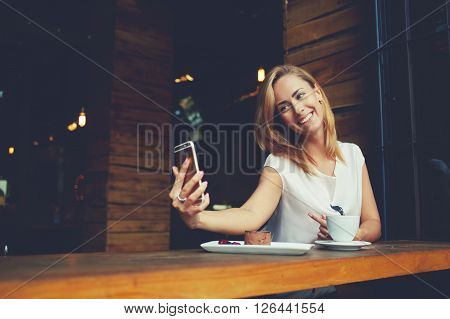 Charming happy hipster girl photographing herself on cell telephone camera while relaxing in modern coffee shop pretty woman with cute smile making self photo on her smart phone during rest in cafe