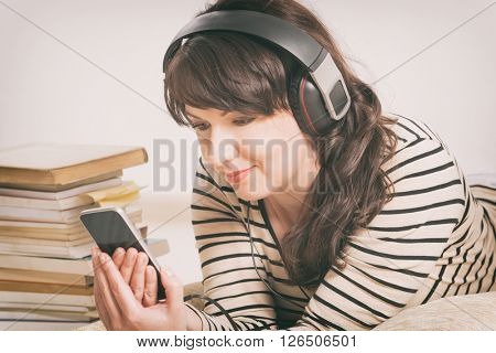Woman listening an audiobook on smartphone sitting between piles of paper books on the floor
