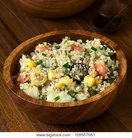 Quinoa salad with sweet corn olive tomato cucumber and chives in wooden bowl photographed on dark wood with natural light (Selective Focus Focus in the middle of the salad)