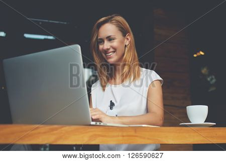 Pretty female student with cute smile keyboarding something on net-book while relaxing after lectures in University beautiful happy woman working on laptop computer during coffee break in cafe bar
