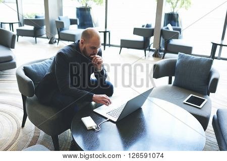 Confident businessman is reading in network via laptop computer customer reviews about his company. Male successful lawyer is studying dealing new client by using net-book while is sitting in cafe