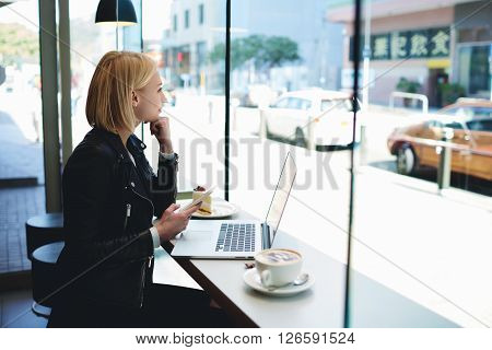 Hipster girl with mobile phone thoughfull look into the window while is sitting in modern coffee shop interior at the table with open laptop computer.Woman freelancer is using cell phone and net-book