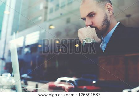 View through the window of a young man professional banker with luxury watch on hand is fulfilling an important banking operations in internet via his portable laptop computer. Double exposition
