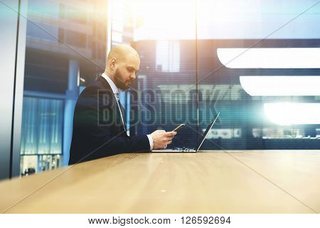 Bearded businessman is calculating the income of the firm last month by using cell telephone and portable net-book. Young male proud banker checking with mobile phone the state of the currency market