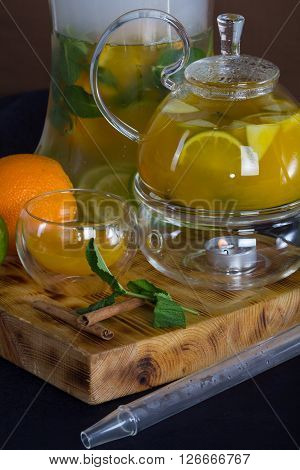 still life in the yellow colors of fruit tea in glass teapot , shisha , mint and orange on a wooden stand .