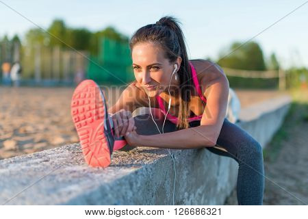 Athletic woman stretching her hamstring, legs exercise training fitness before workout outside on a beach at summer evening with headphones listening music.