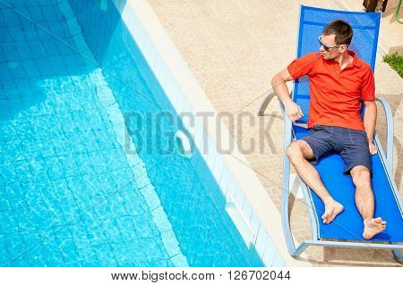 young man relaxing on the chaise-longue near the pool. man wearing in red polo shirt and sunglass