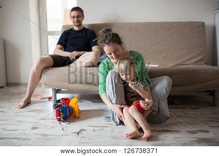 Cheerful mother is teaching toddler son father near. She is holding him and sitting on flooring. The mom is looking at her child with love casual lifestyle photo series in real life interior