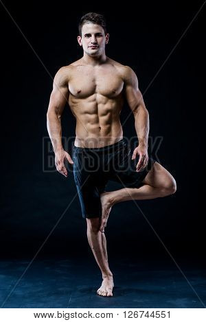 Muscular man balancing on the one leg feeling relaxed on dark background