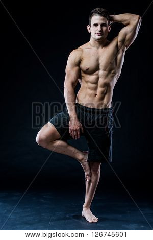 Muscular man balancing on the one leg feeling relaxed on dark background