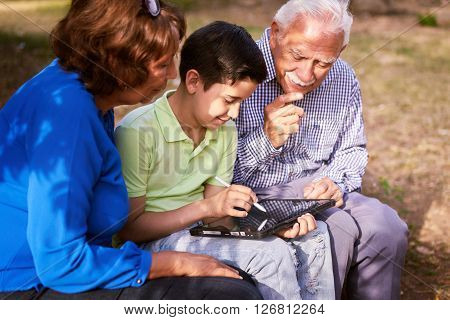 Grandparents educating grandson: Senior woman and old man spending time with their grandchild in park. The old people help the boy studying and doing school homework. The kid holds a tablet pc