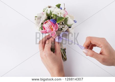 Two hands are making a bow from a violet ribbon in beautiful spring bouquet from pink tulips violet grape hyacinths white anemones violet veronica and white buttercups on white background
