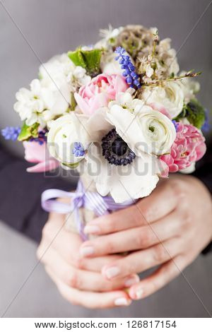 Woman in grey dress and black shirt is holding in her hands a spring bouquet from pink tulips violet grape hyacinths white anemones violet veronica and white buttercup with violet ribbon