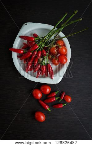 Top view on cherry tomatoes and mini red hot chili peppers in old plate on dark wooden table. Dark food photography with bunch of red chili and tomatoes.