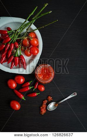 Glass jar of homemade tomato sauce with fresh ingredients on dark wooden background. Natural homemade sauce of tomatoes, peppers and vegetables. Fresh Homemade Salsa Dip. Chilli jam. Top view.