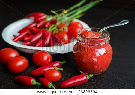 Glass jar of homemade tomato sauce with fresh ingredients on dark wooden background. Tomato ketchup sauce with cherry tomatoes and mini red hot chili peppers. Selective focus.