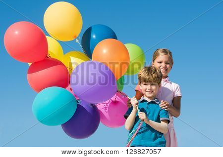 Cute boy and girl playing with colorful balloons outdoor. Happy smiling children looking at camera with balloons. Happy sister and brother having fun with balloons with blue sky in background.