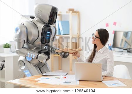 My best helper. Delighted charming positive smiling girl sitting at the table and working on the laptop while getting package from the robot