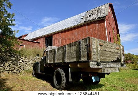 A deteriorating huge red barn has an old black truck with a wood box parked in front.