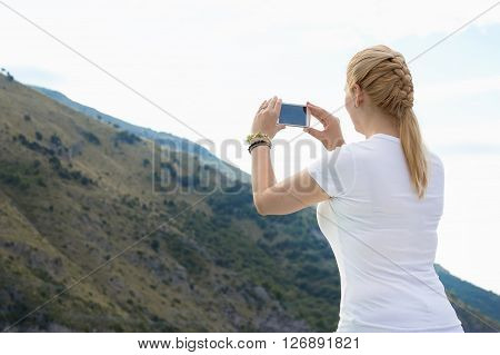 Young beautiful blonde woman doing a picture with your mobile phone. A woman is standing against a background of say and mountains she is a tourist. Photograph mobile phone.