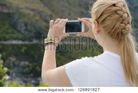 Young beautiful blonde woman doing a picture with your mobile phone. A woman is standing against a background of mountains she is a tourist. Photograph mobile phone.