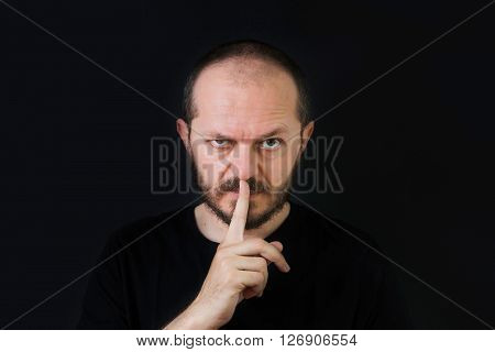 Serious man with beard and mustaches on black background in low key, making silence gesture, pst, shh