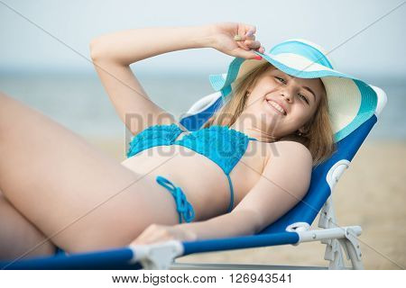 Young lady sunbathing on a beach. Beautiful woman posing at the summer sand beach. Outdoor summer portrait of pretty sport style woman in blue bikini. Ocean sea coast. Beautiful fit tan girl. Sexy slim model caucasian ethnicity outdoors.