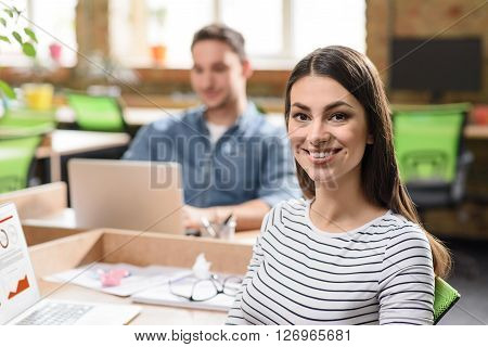 Positivity in mind. Cheerful charming delighted woman smiling and sitting at the table  while her colleague sitting in the background  and working on the laptop