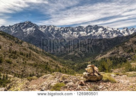 Small stone cairn in the mountains in Corsica with a pine tree covered valley and snow capped Asco mountains and Monte Cinto in the distance