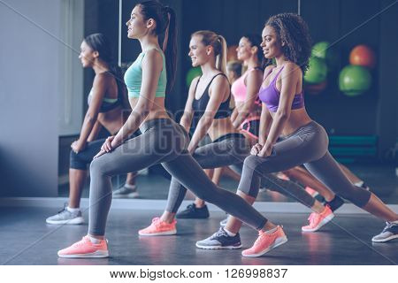 Stretching with smile. Side view of beautiful young women with perfect bodies in sportswear exercising with smile at gym