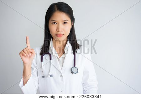 Young Asian Female Physician Pointing Finger Up. Serious Health Worker Showing Warning Gesture. Medi