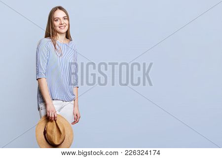 Horizontal Shot Of Happy Female In Blouse And White Trousers, Holds Straw Hat, Being Happy To Be Pho