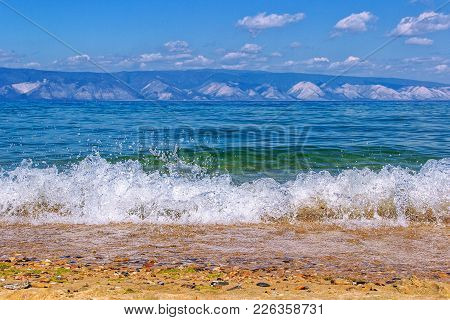Landscape With The Image Of Running Waves On The Coast Of Lake Baikal And Mountains In The Backgroun