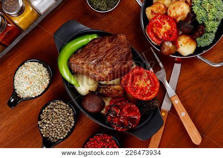fresh roast beef meat rib eye steak with tomatoes,potatoes, hot chili pepper gern and pink peppercorn saucer, cutlery, set of spice, and red wine glass on light walnut table
